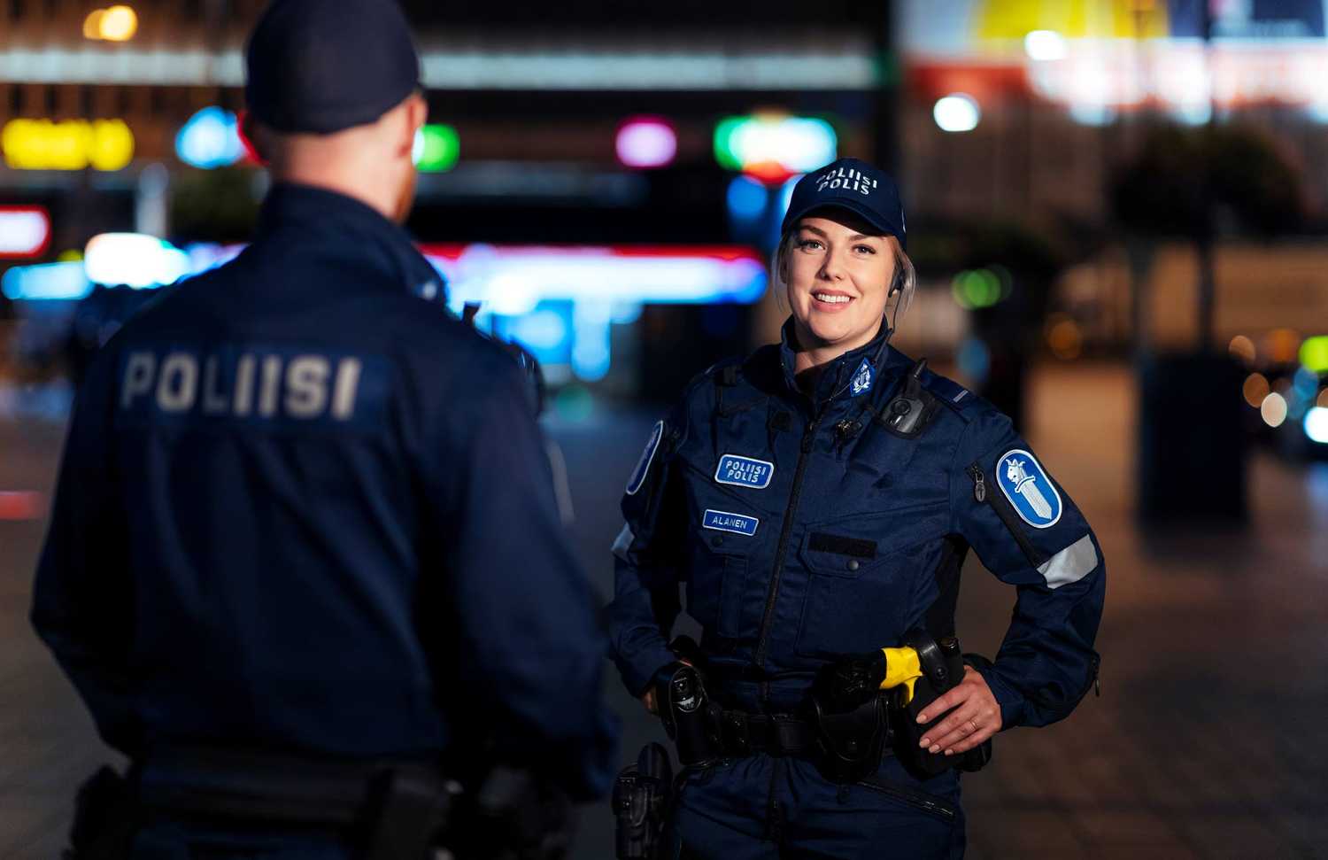 Two police officers in blue uniforms, the female police officer facing the camera, the red-bearded male police officer with his back to the camera. In the background, the city's colorful advertising lights.