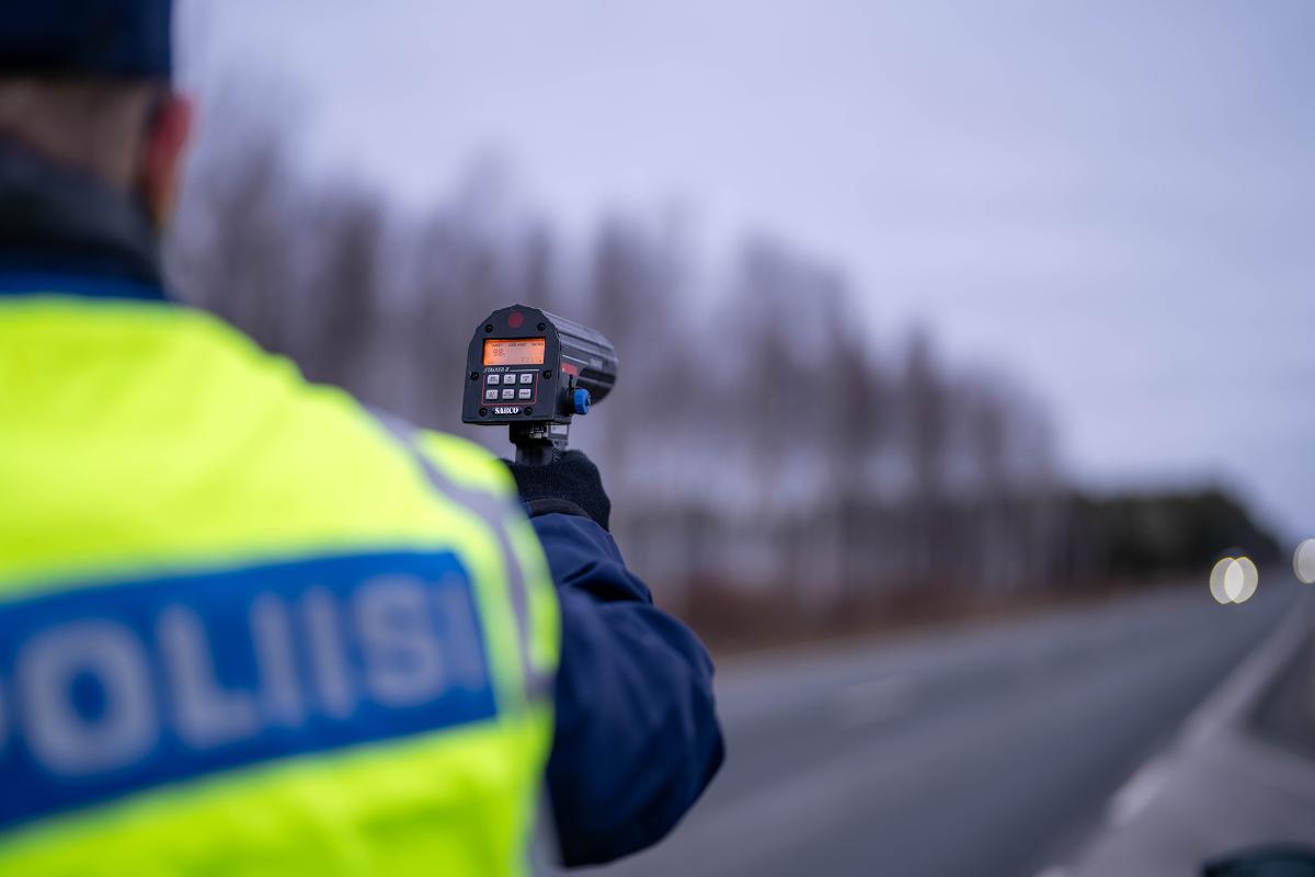 Kuva poliisista mittaamassa tutkalla lähestyvän auton nopeutta. Poliisilla on yllään keltainen heijastinliivi.