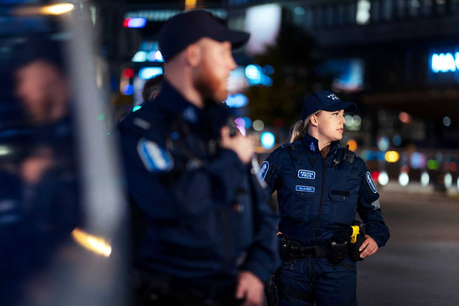 Poliisipartio seisoo poliisiauton vierellä. Toinen poliiseista puhuu puhelimeen. Taustalla näkyvät kaupungin mainosvalot.