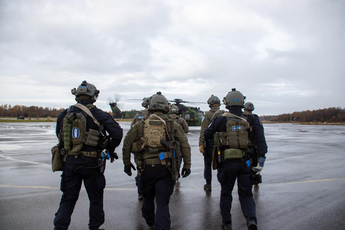 Etualalla viisi karhun erikoisoperaattoria, jotka kävelevät edessa olevaa Puolustusvoimien helikopteria kohti.
