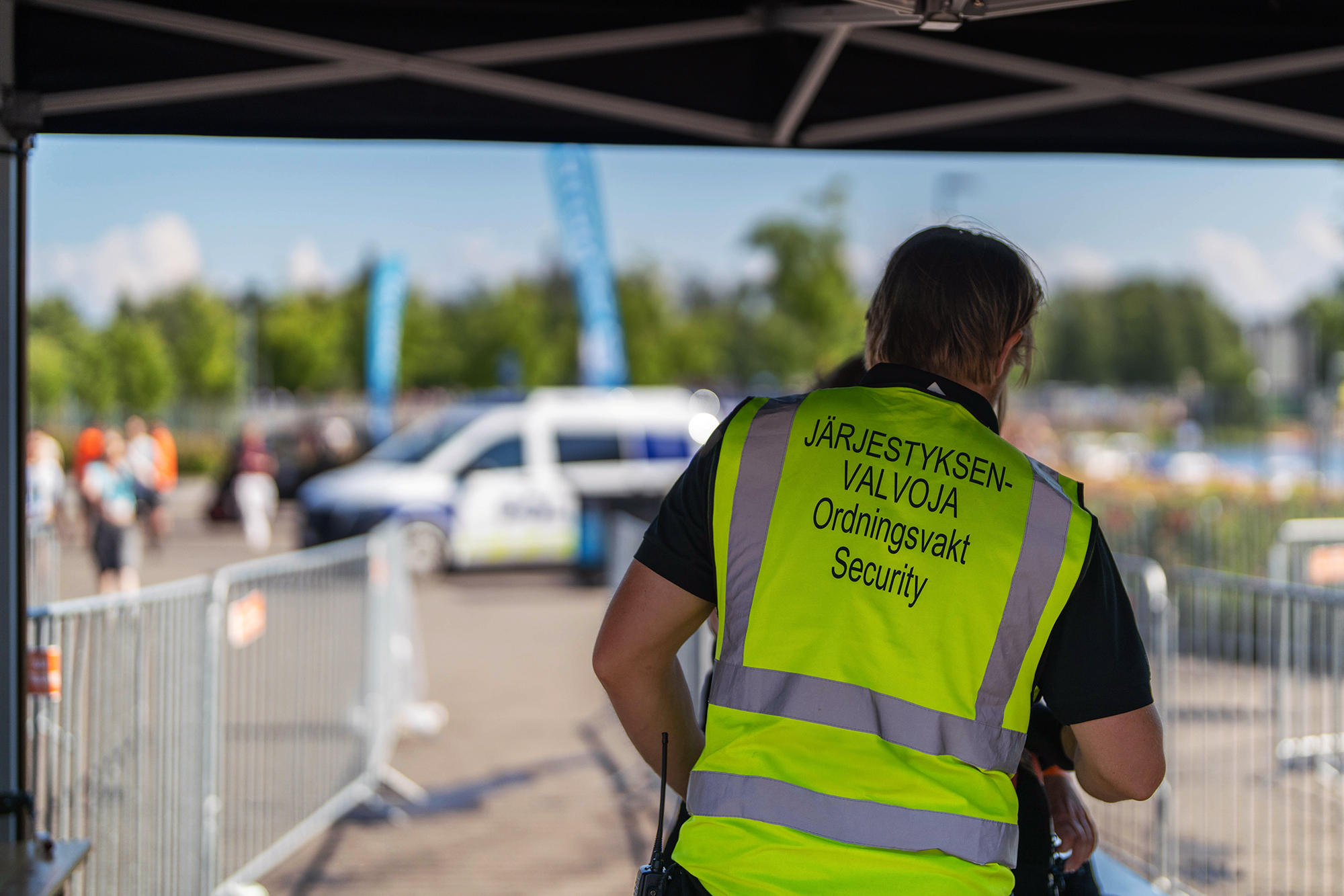Järjestyksenvalvoja tarkastaa tapahtumaan tulevaa vierailijaa portilla. Taustalla poliisiauto sumeana.