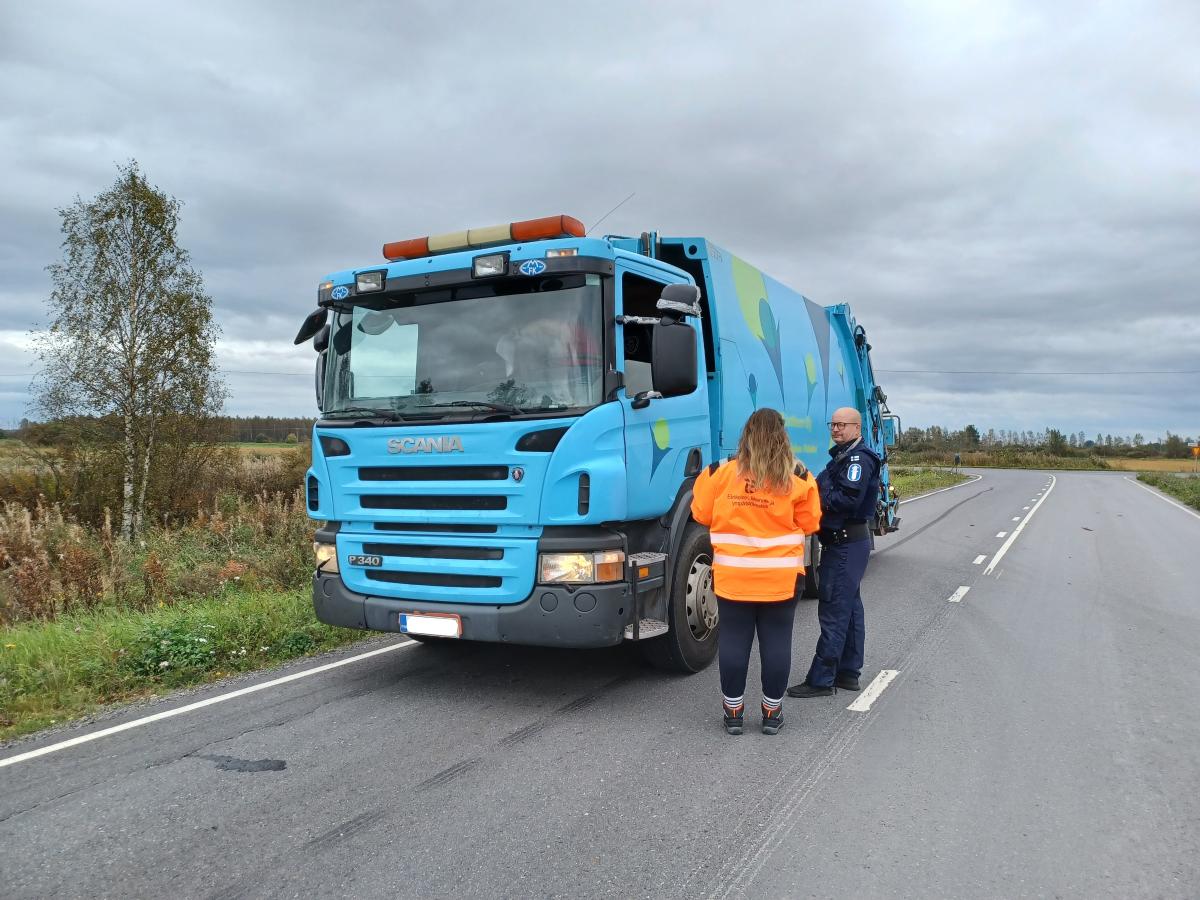 Jäteauto tien reunassa. Edustalla kuljettaja ja poliisi keskustelevat. Kuljettajalla on oranssi takki.