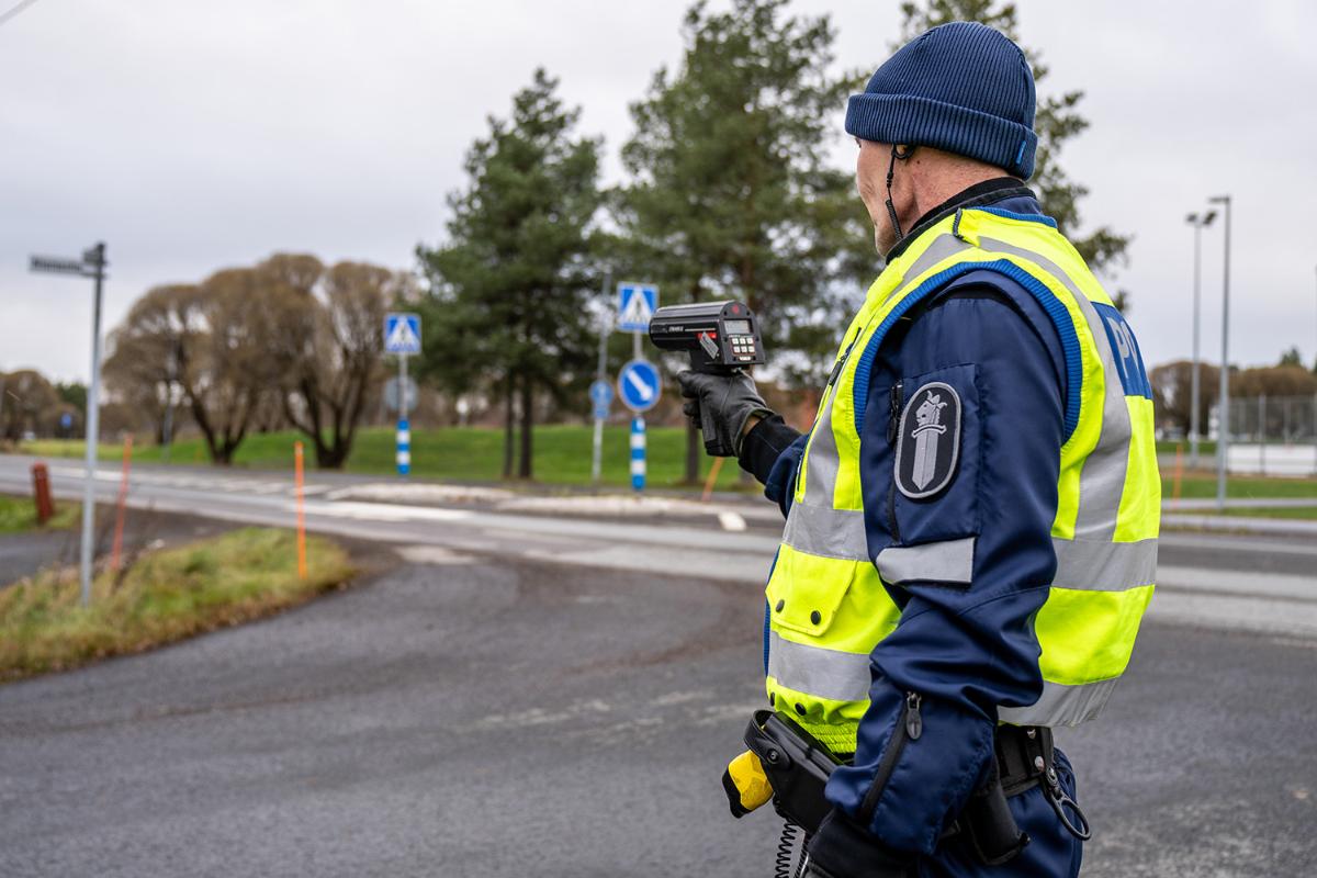 Poliisi seisoo tienristeyksessä ja mittaa ajoneuvon nopeutta liikenteessä nopeusmittarilla. Poliisilla on yllään keltainen huomioliivi ja päässä pipo.
