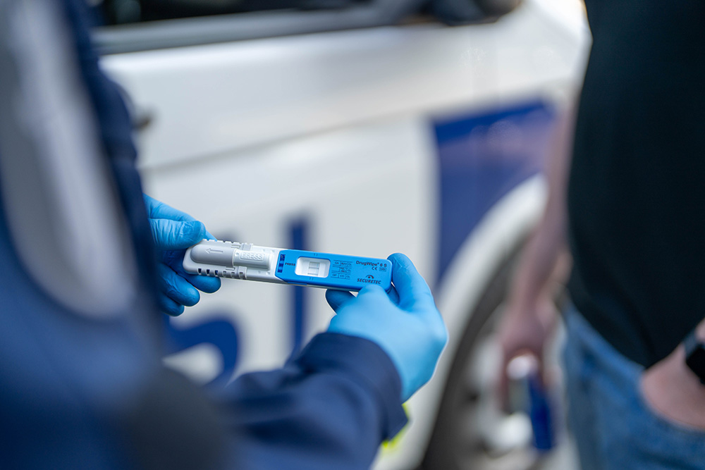 Poliisi pitää käsissään huumepikatestiä. Taustalla poliisiauto.