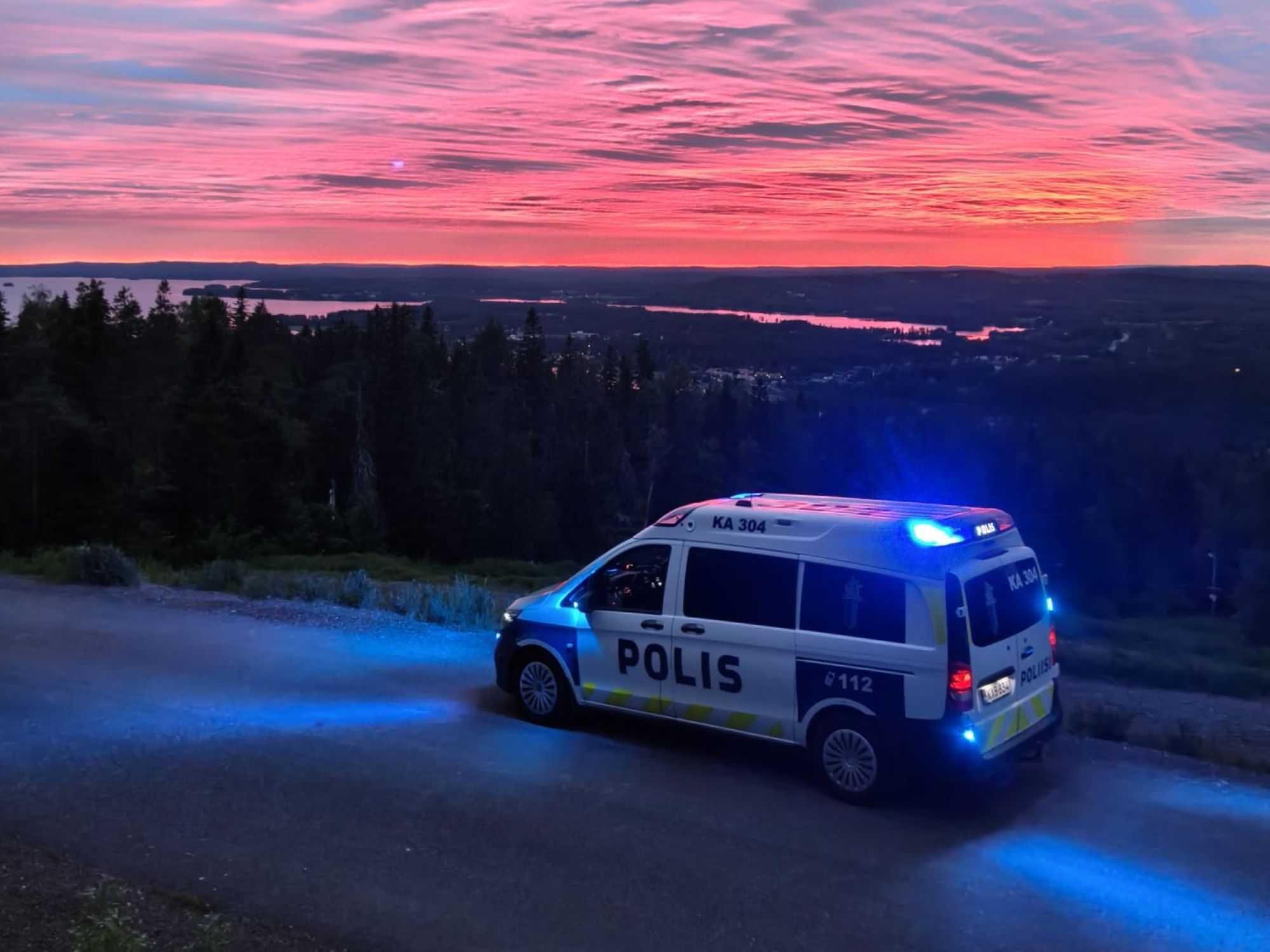 Poliisiauto punaisena hohkaavassa auringonlaskussa Vuokatinvaaran huipulla Sotkamossa. Autossa vilkkuu hälytysvalot.