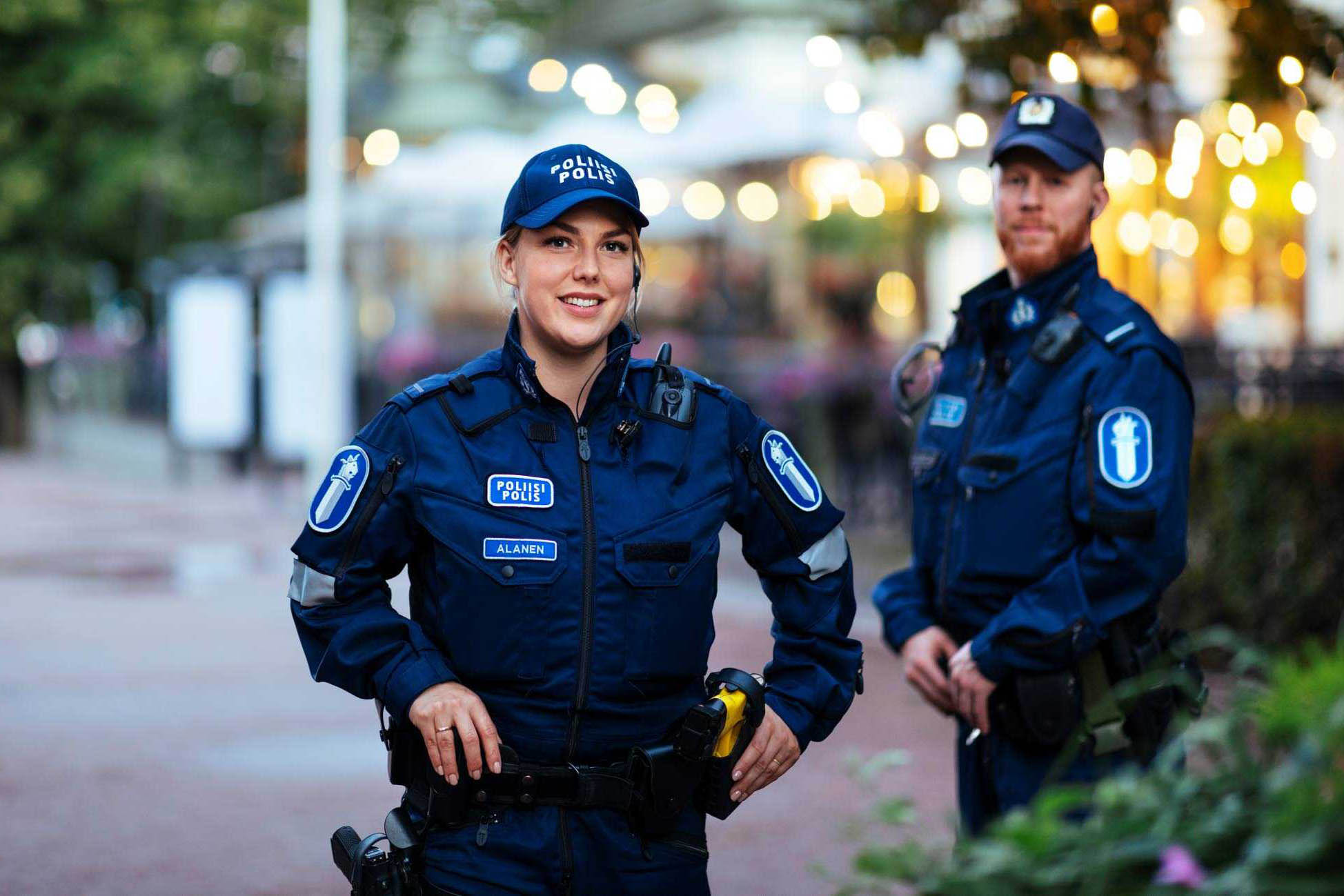 Kaksi poliisia kasvot kuvaajaan päin, etualalla naispoliisi ja taaempana seisoo punapartainen miespoliisi. Molemmilla päällä sininen poliisin kenttähaalari varusteineen ja päässä poliisin lippalakit. Taustalla kasvillisuutta ja kaupungin valoja.