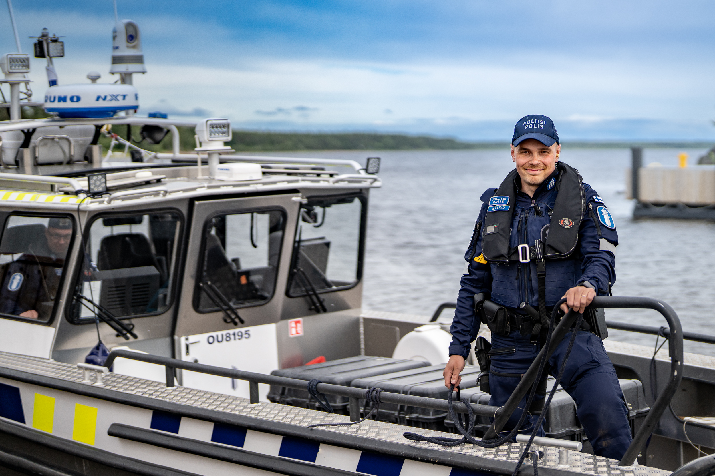 Hymyilevä poliisi seisoo partioveneen keulassa veneen rantautuessa. Taustalla toinen poliisi ohjaa venettä ja näkyy sumeasti. Sää on pilvinen.