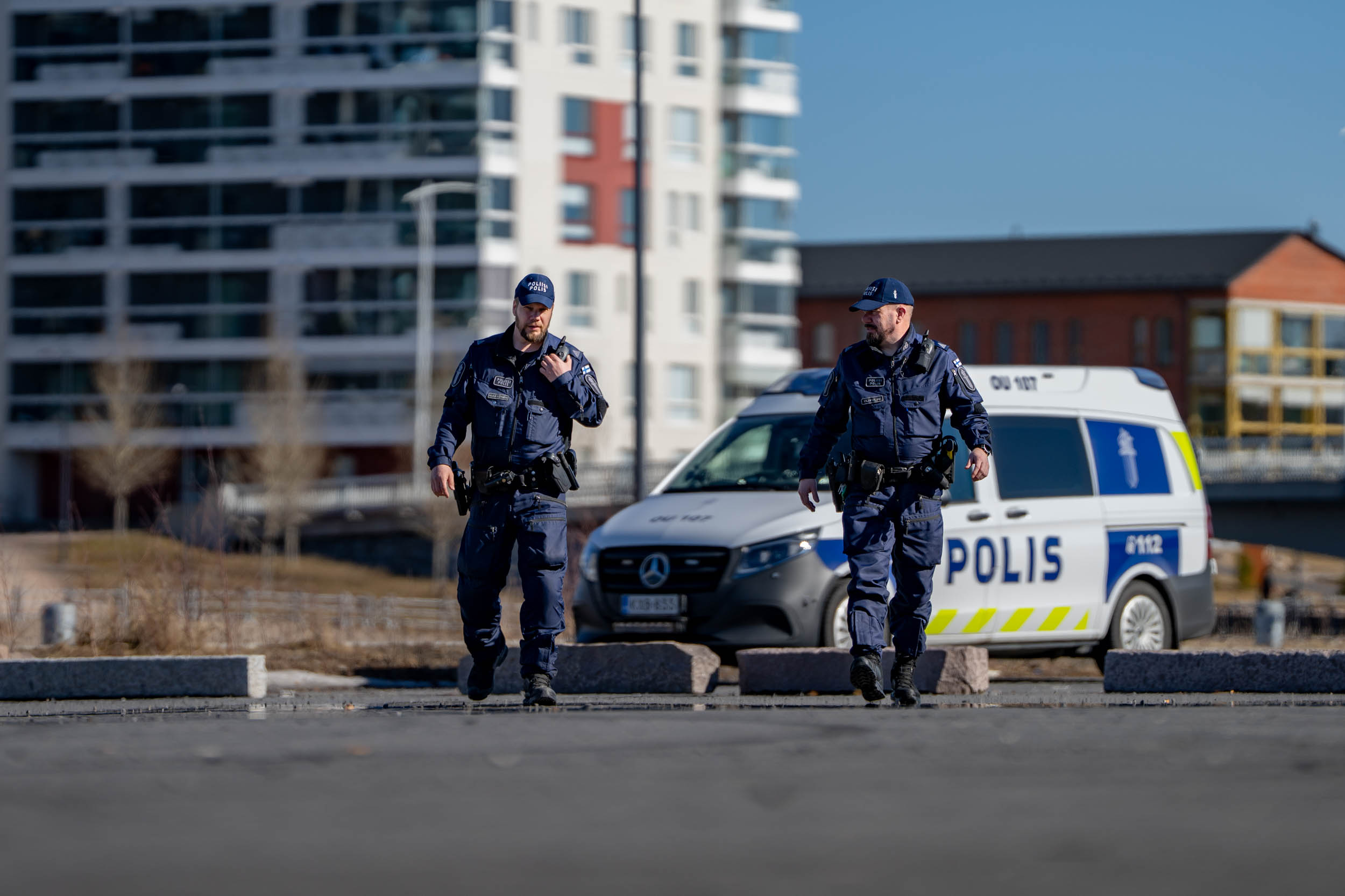 Två poliser promenerar i Toppilansalmi i Oulu i vårväder. Himlen är klar och solen skiner. En av poliserna pratar i radion. I bakgrunden syns en polisbil och flerbostadshus.