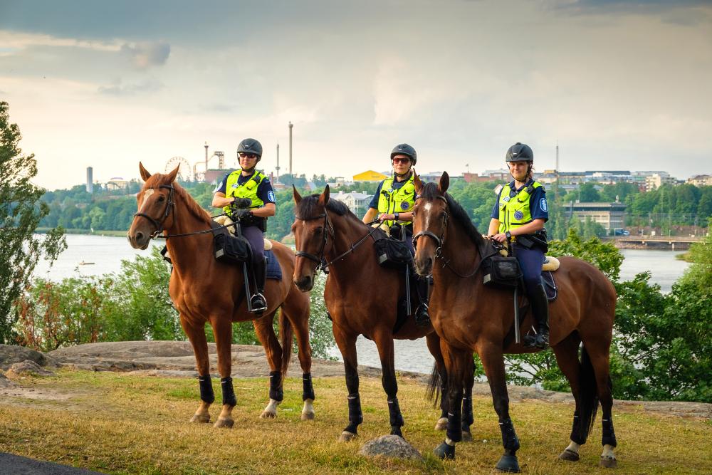 Three mounted police officers side by side with Helsinki’s Töölönlahti bay in the background on a bright summer day.