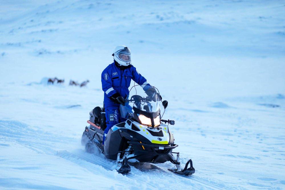 Poliisi ajaa talvivarusteissa moottorikelkalla keskellä lumista maisemaa. Taustalla häämöttää poroja.