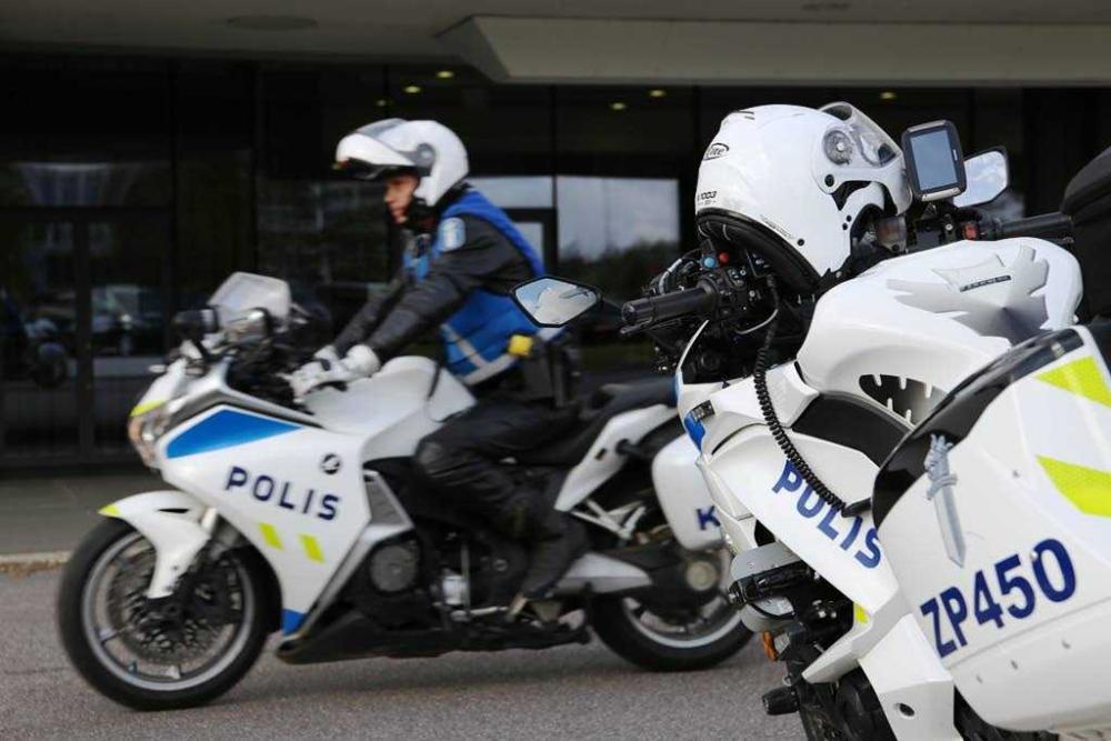 Två motorcykelpoliser i eskortkörning. I förgrunden en motorcykel utan förare och rakt fram ses en motorcykelpolis som är redo att köra iväg. 