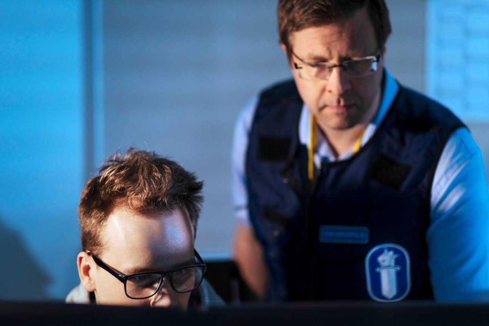 Two analysts looking at a computer screen, one wearing an overt police vest.