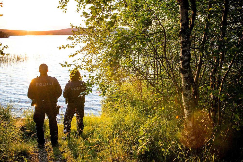 On the shore of the lake, two police officers in overalls look out over the lake. The sun is setting, surrounded by summer nature. 