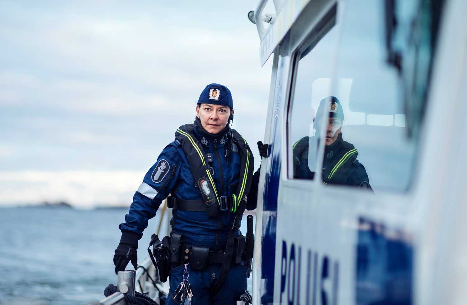 Båtpolisen står på polisbåtens däck, lutad mot räcket, bredvid kan man se texten poliisi på sidan av båten.