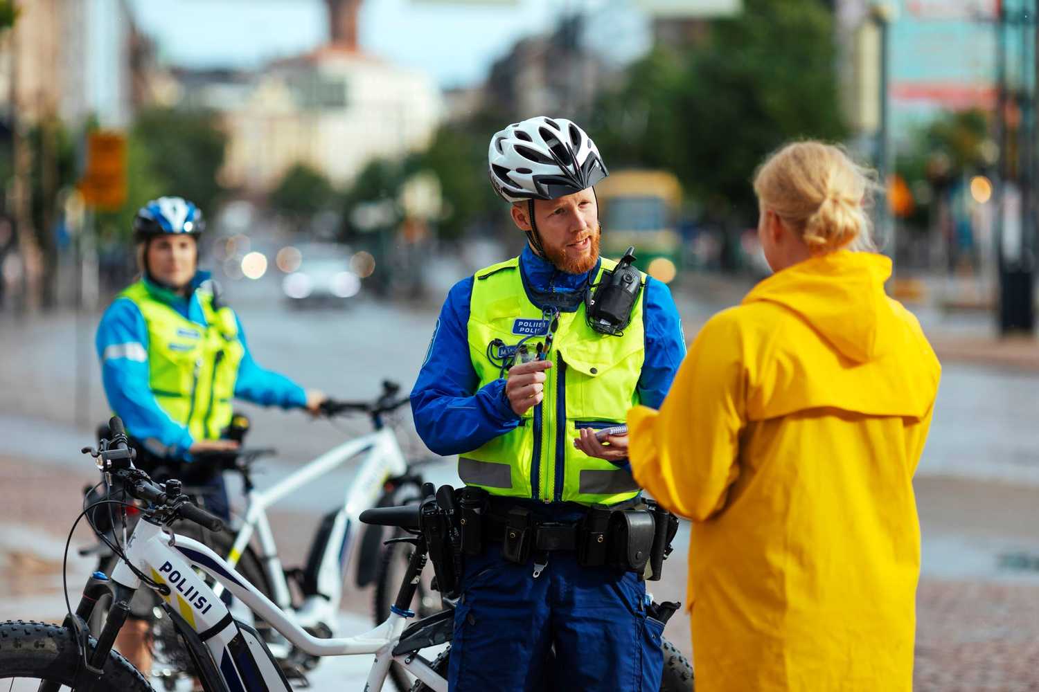 Polkupyöräpoliisi puhuttaa keltaiseen takkiin pukeutunutta asiakasta. Poliisilla on kädessään kynä ja lehtiö. Taustalla toinen poliisi nojaa pyöräänsä.