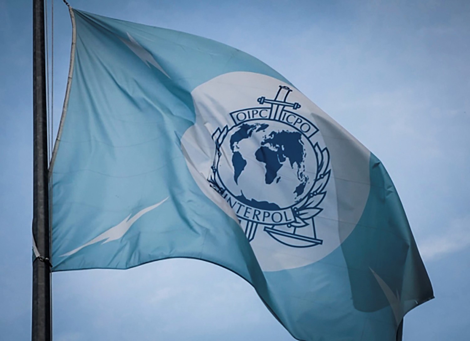 Interpol blue and white flag in salo. It has a light blue background with an emblem in the center and four lightning bolts symbolizing telecommunications and speed in policing.