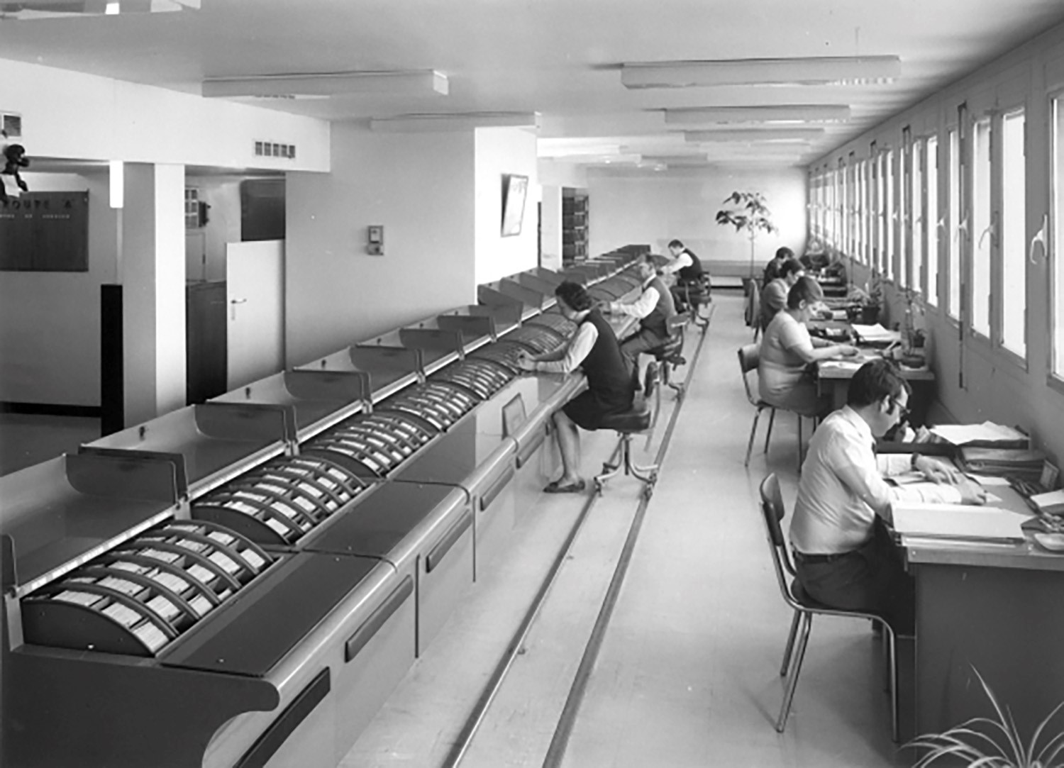 The black-and-white picture shows men and women in the hall at desks.