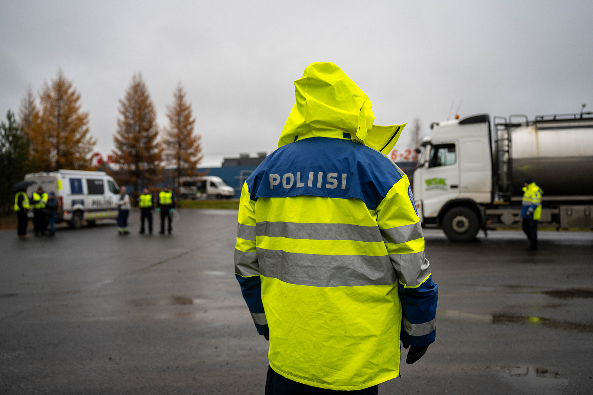 Poliisi on raskaan liikenteen valvonnassa Pohjois-Pohjanmaalla Iissä syksyllä. Poliisi on kuvattu selästä ja käyttää keltaista sadetakkia sateisessa säässä. Taustalla näkyy kuorma-auto sekä Tullin edustajia ja poliiseja sumeana.
