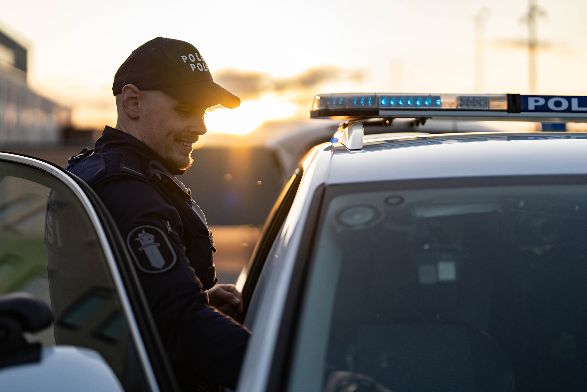 A police officer stands next to a Skoda Kodiaq police car, smiling toward the vehicle. Oulu main police station in Ruskonselkä and a sunset are visible in the background.
