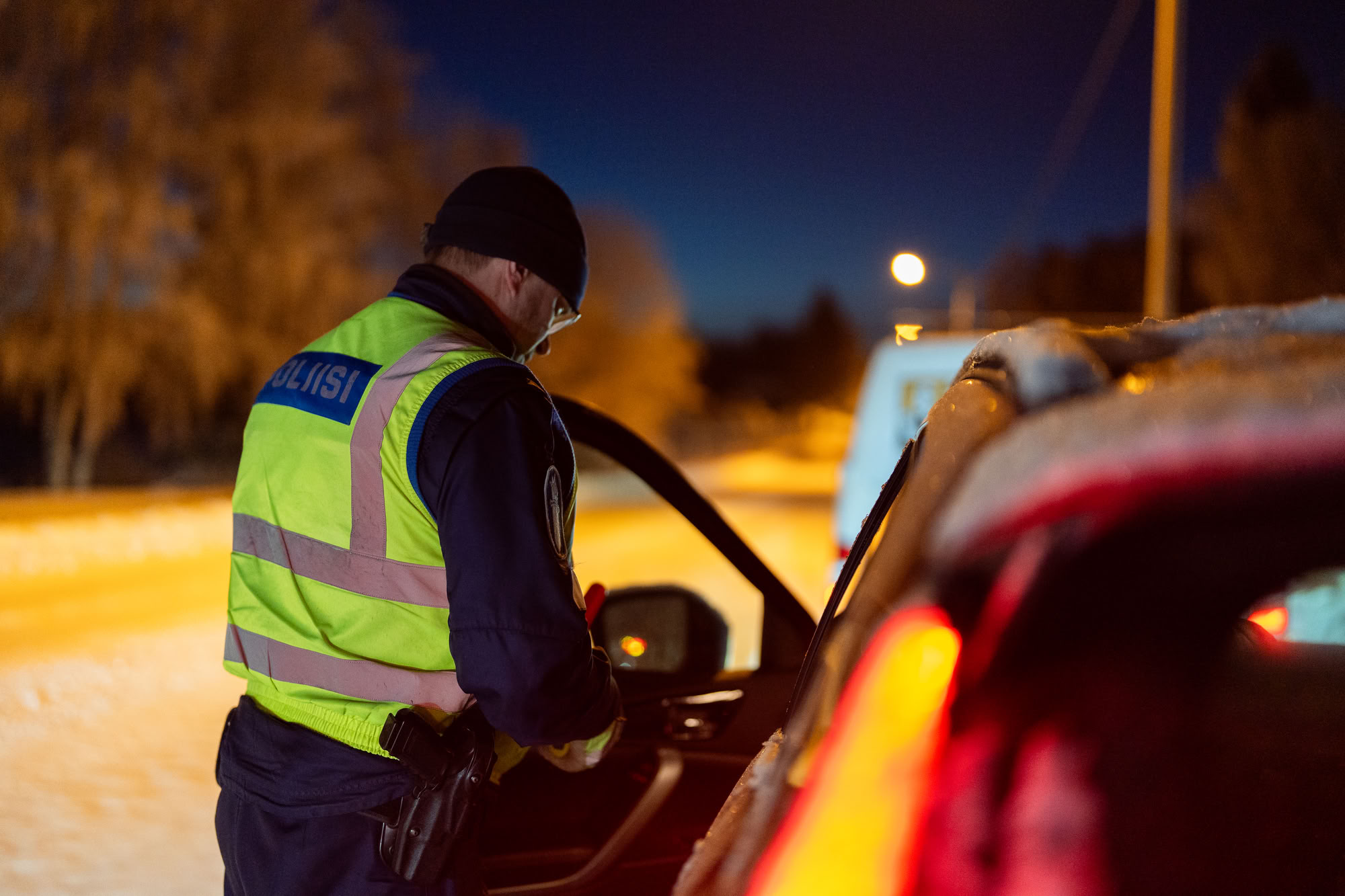 A police officer wearing a high-visibility vest has stopped a car and conducts a breathalyzer test early in the morning. The surroundings are lit by streetlights.