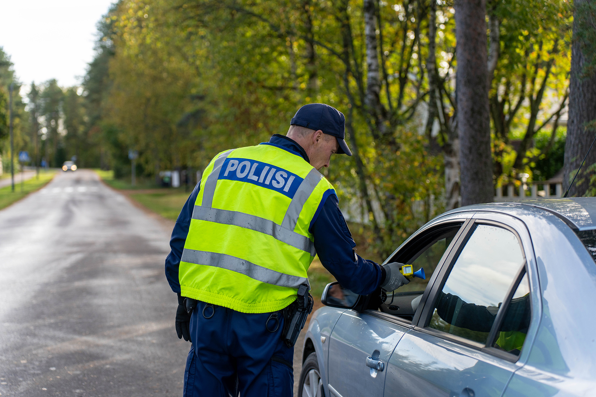 A police officer wearing a high-visibility vest conducts a breathalyzer test on a driver in front of a daycare center in Ylivieska during autumn. The road continues in the background.