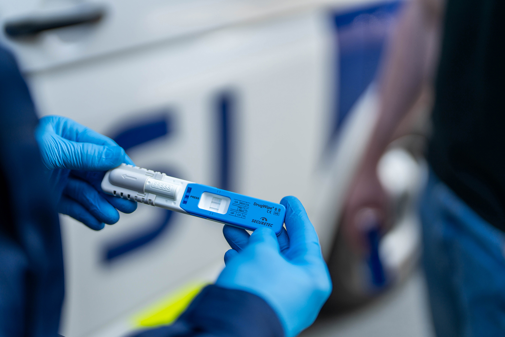 A police officer performs a rapid drug test while wearing blue protective gloves. The side of a police car and a related person appear blurred in the background.