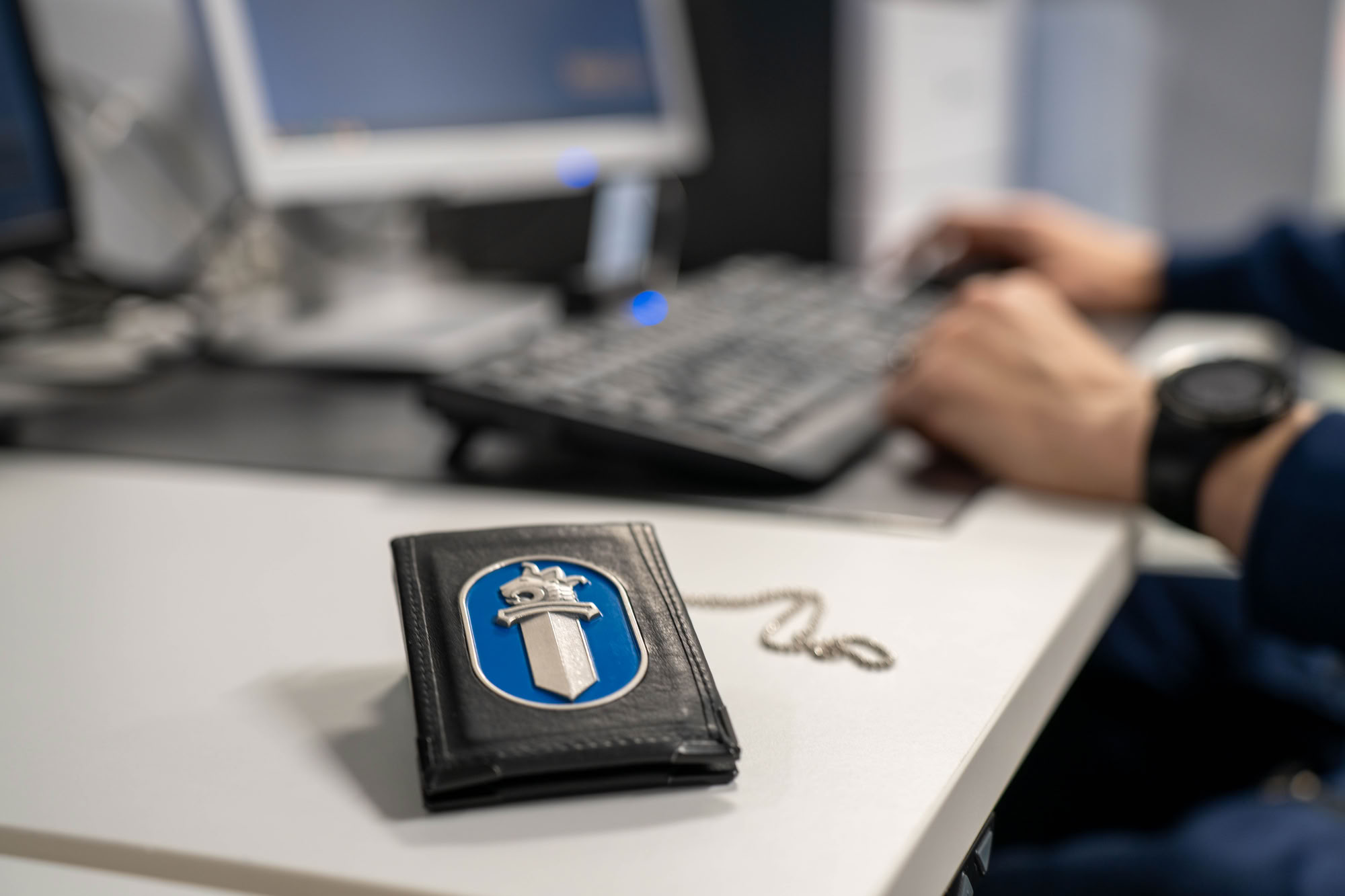 Poliisin virkamerkkikotelo on etualalla. Taustalla poliisi työskentelee tietokoneella.