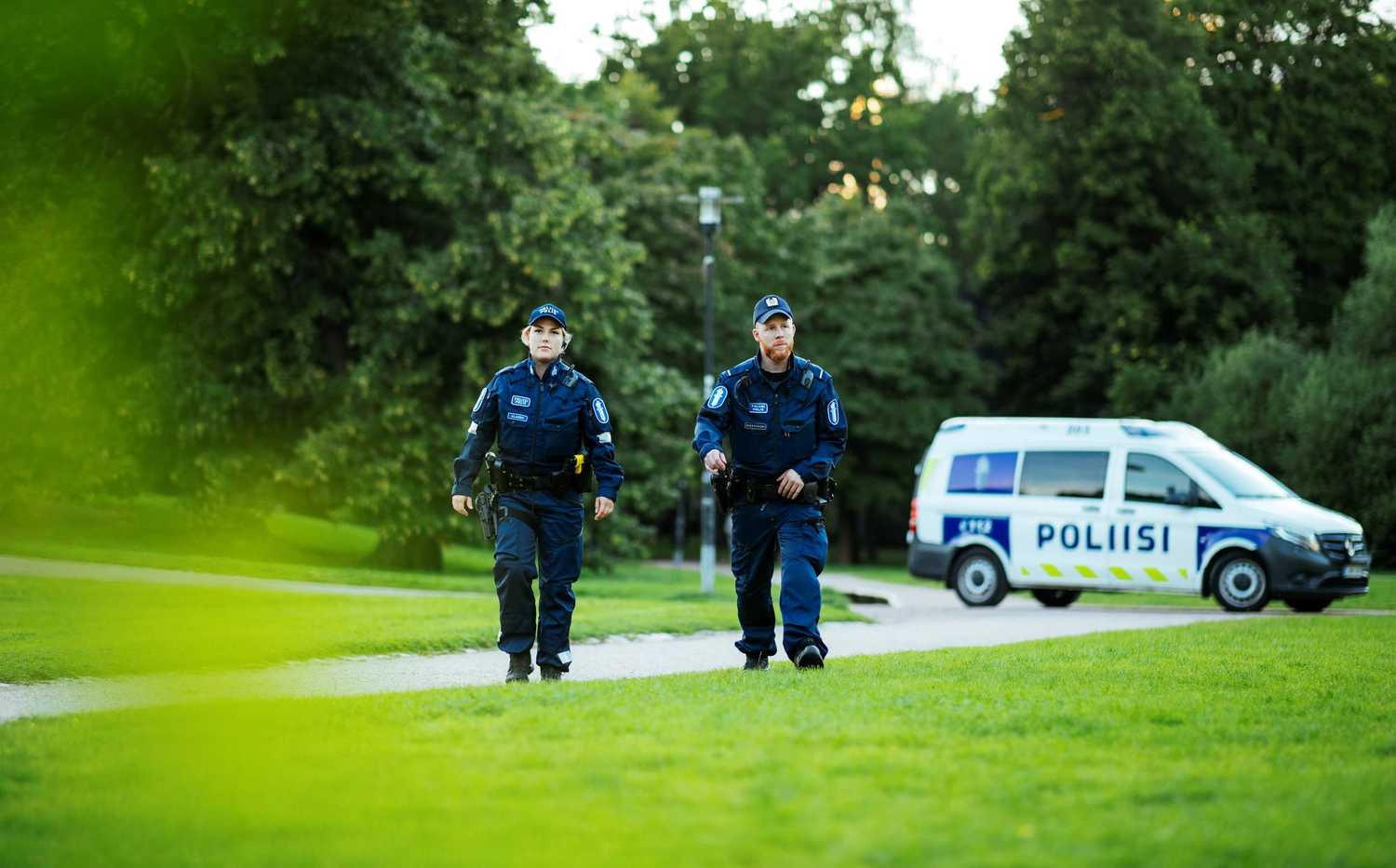 Two police officers in uniform walking in a park at summer. A police car in the backround.