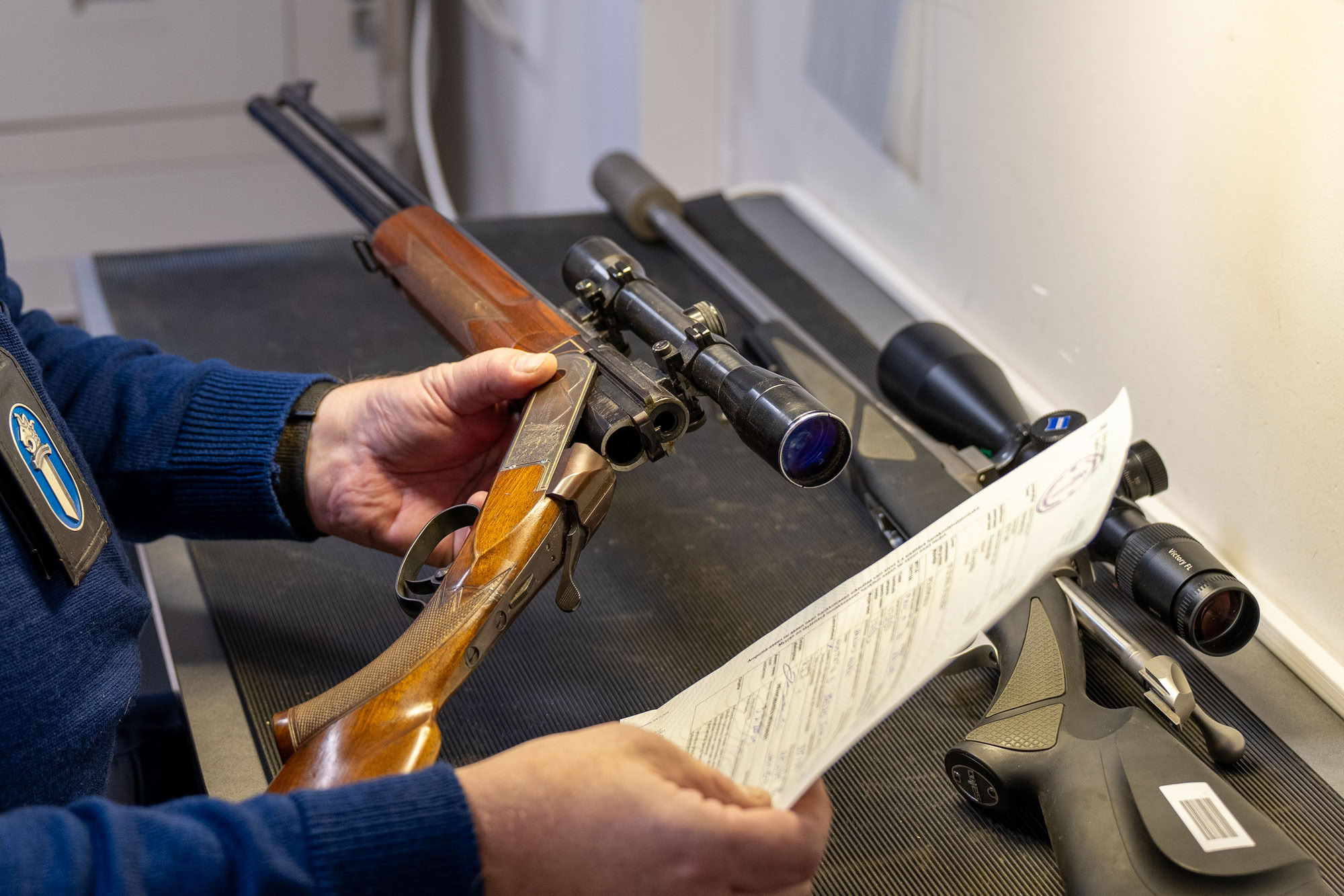 Asetarkastaja vertailee lomakkeessa olevia aseen tietoja käsissään olevaan aseeseen. Kädet, lomake ja kaksi asetta näkyvät.