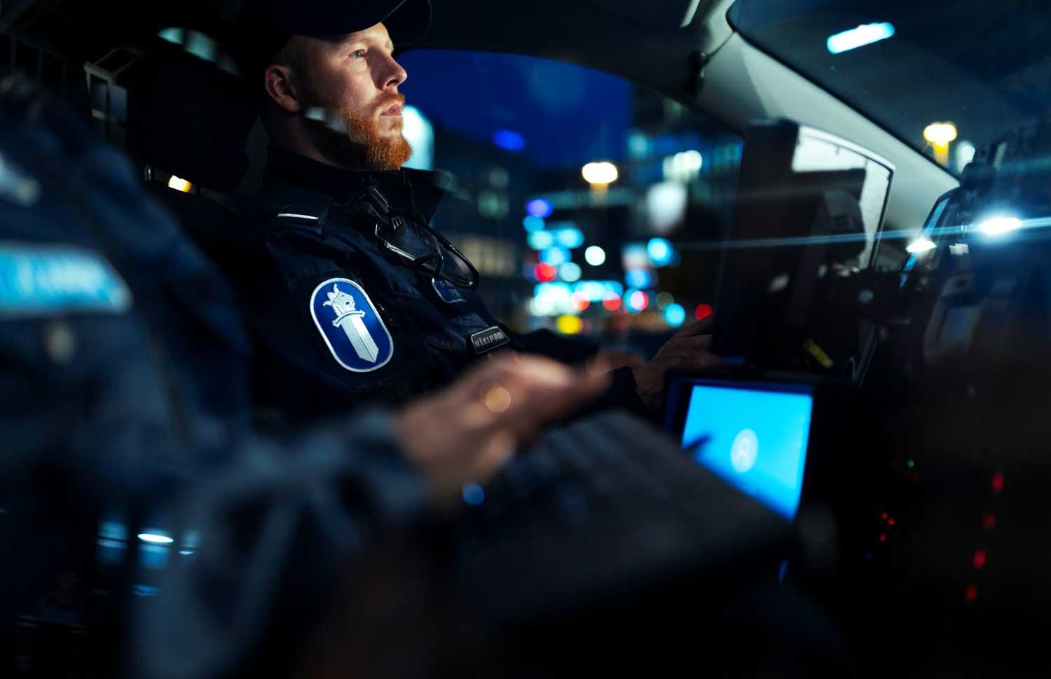 Två poliser sitter i en polisbil. Helsingfors stadsljus syns i bakgrunden. 