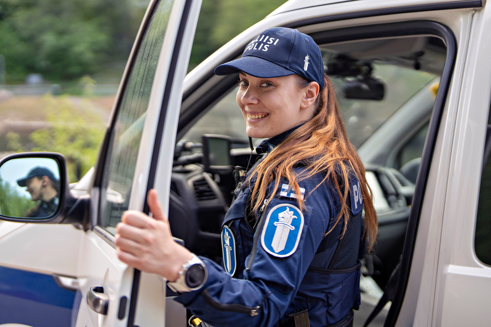 En polis ser leende mot kameran. En polisbil syns i bakgrunden.