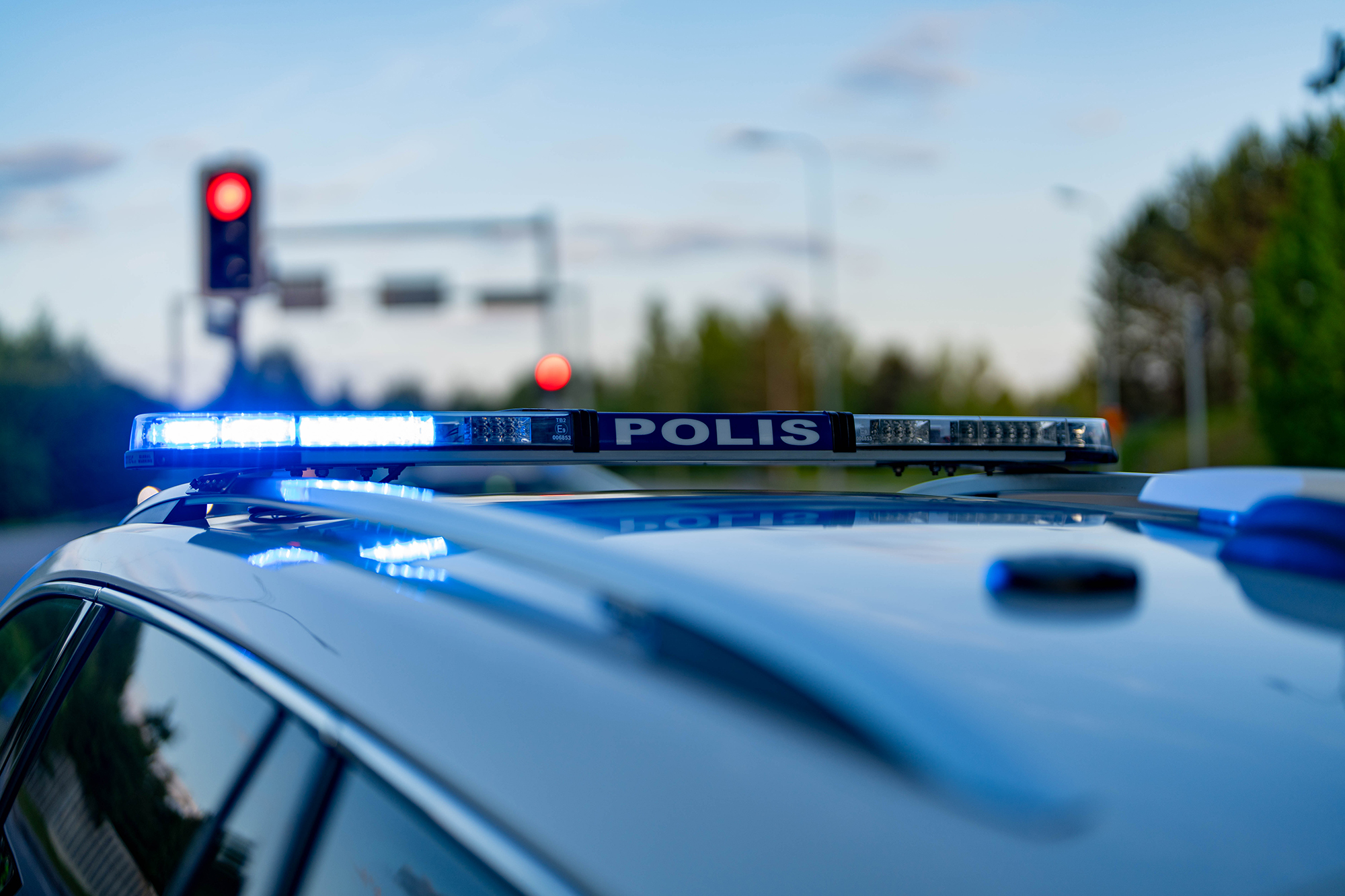Emergency lights flash on top of a police car, with red traffic lights visible in the background.