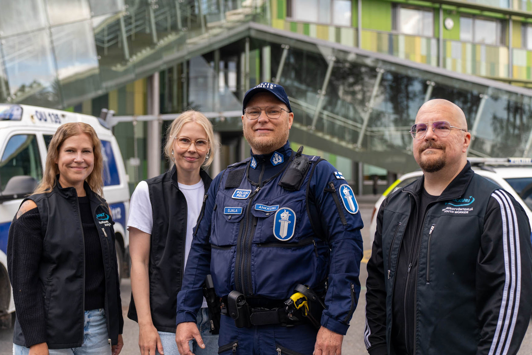 Fyra medlemmar av Oulus Anchor-team — experter från polis, social-, hälso- och ungdomssektorn — står framför en skola med två polis­bilar i bakgrunden.