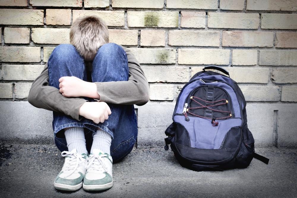 En skolelev med huvudet mot knäna med en ryggsäck framför en tegelmur. 