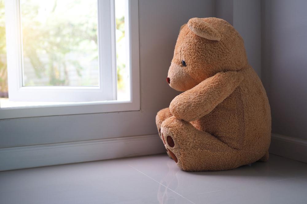 A teddy bear sitting on a window sill looking out. 