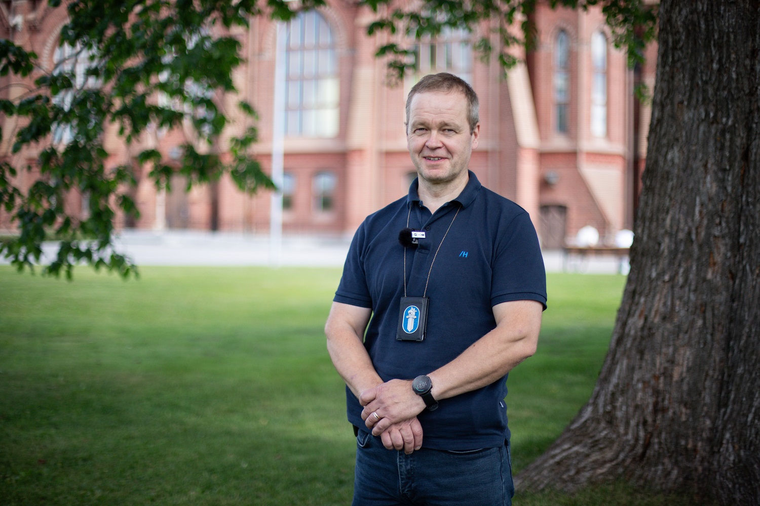 Detective Sergeant Ismo Venäläinen in a blue shirt stands at the base of a large deciduous tree, a police badge with a blue and white police sword lion hanging around his neck. In the background you can see a grassy field and a large reddish building.