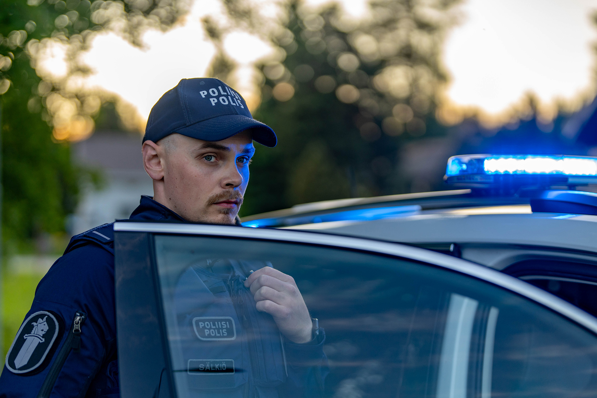 A police officer speaks into a radio on a summer evening, with the patrol car’s emergency lights flashing in the background.