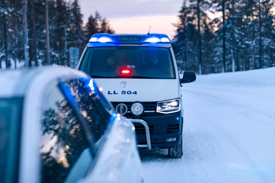 Police car in a winter landscape with its emergency lights on. In the foreground, a stopped car.