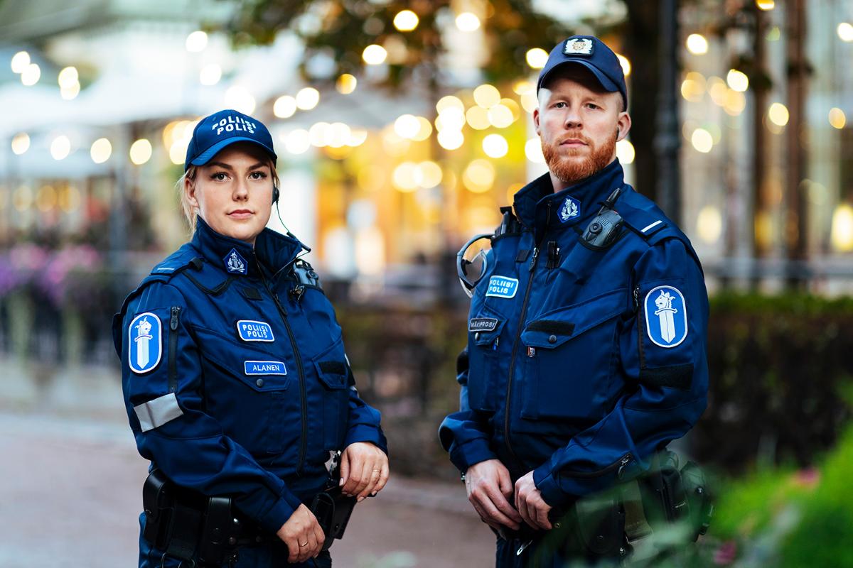 Två uniformerade poliser som poserar i stadsmiljö, namnen Alanen och Mäkiprosi står på namnskyltarna. I bakgrunden butikerna lampor och buskar.