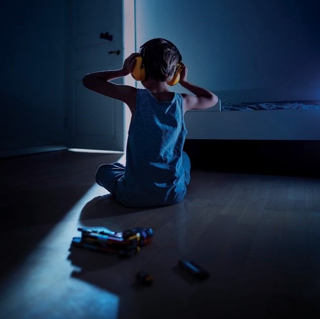 Ett barn sitter i ett mörkt rum med dörren på glänt. Barnet bär hörselskydd.