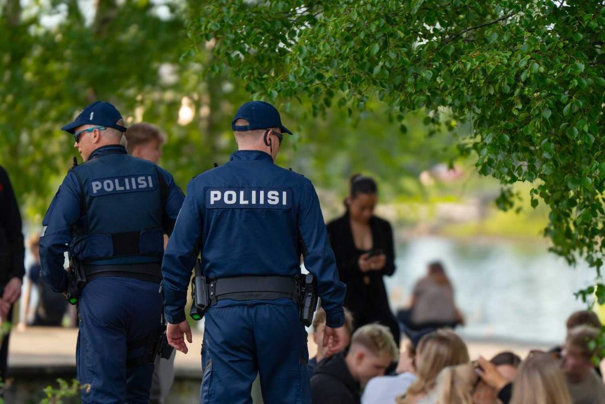 Kaksi poliisia kävelee puistossa, jossa ihmisiä istuu nurmikolla.