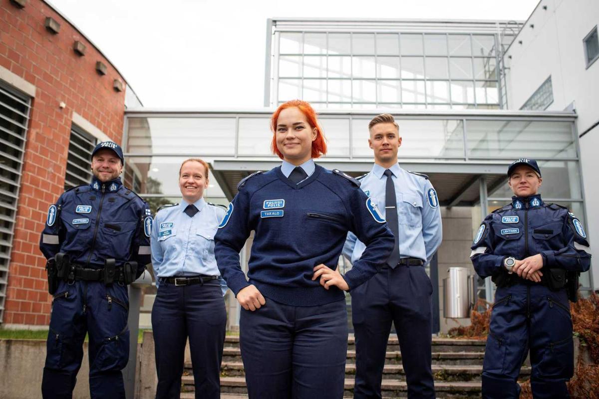 Five police officers stand in front of a building. A red-haired female police officer is in the foreground.