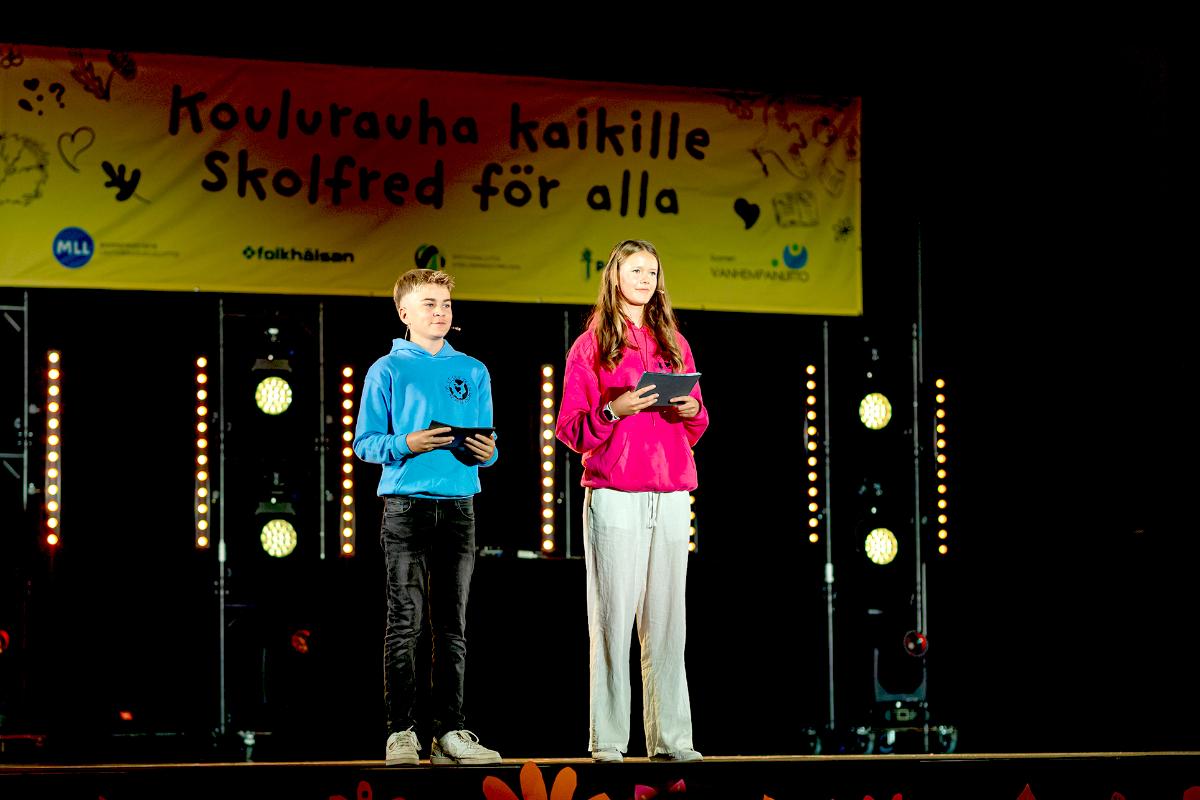 Tyttö ja poika seisovat esiintymislavalla. Taustalla teksti Koulurauha kaikille - Skolfred för alla.