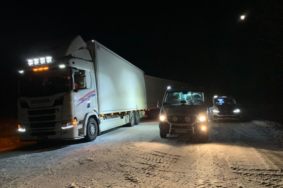 Iso ajoneuvo pysäytettynä lumisella tiellä yöllä. Lisäksi kaksi poliisiautoa.