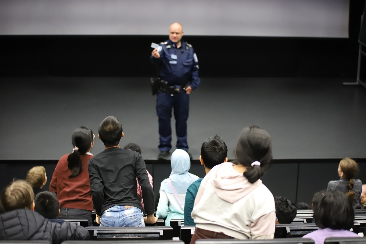 Polisen står på scenen i en skola. Barnen, som sitter på läktaren lutar sig mot honom för att se material i polisens hand.