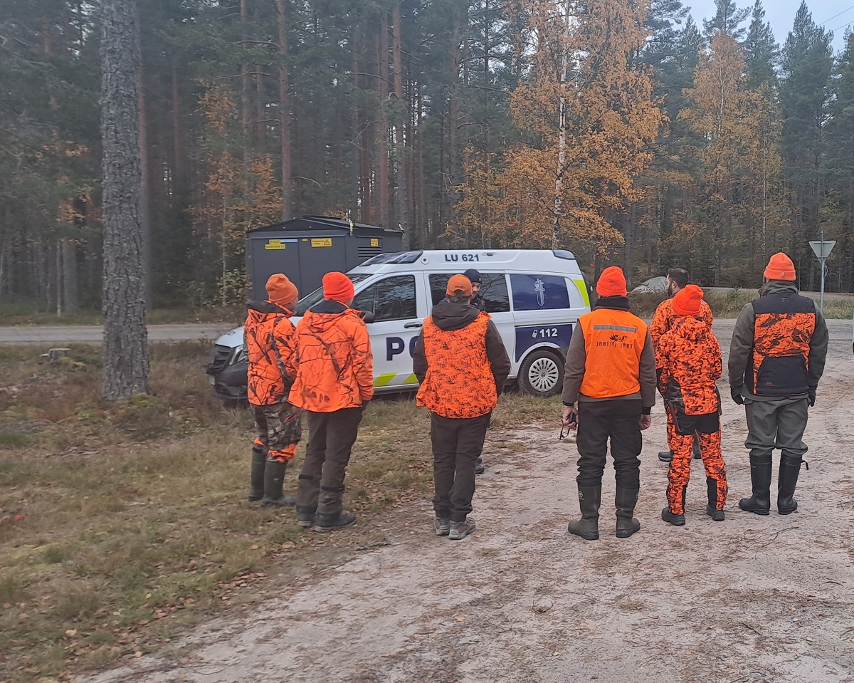 Joukko metsästäjiä seisoo parkkipaikalla ja juttelee poliisiauton edessä seisovan poliisin kanssa.