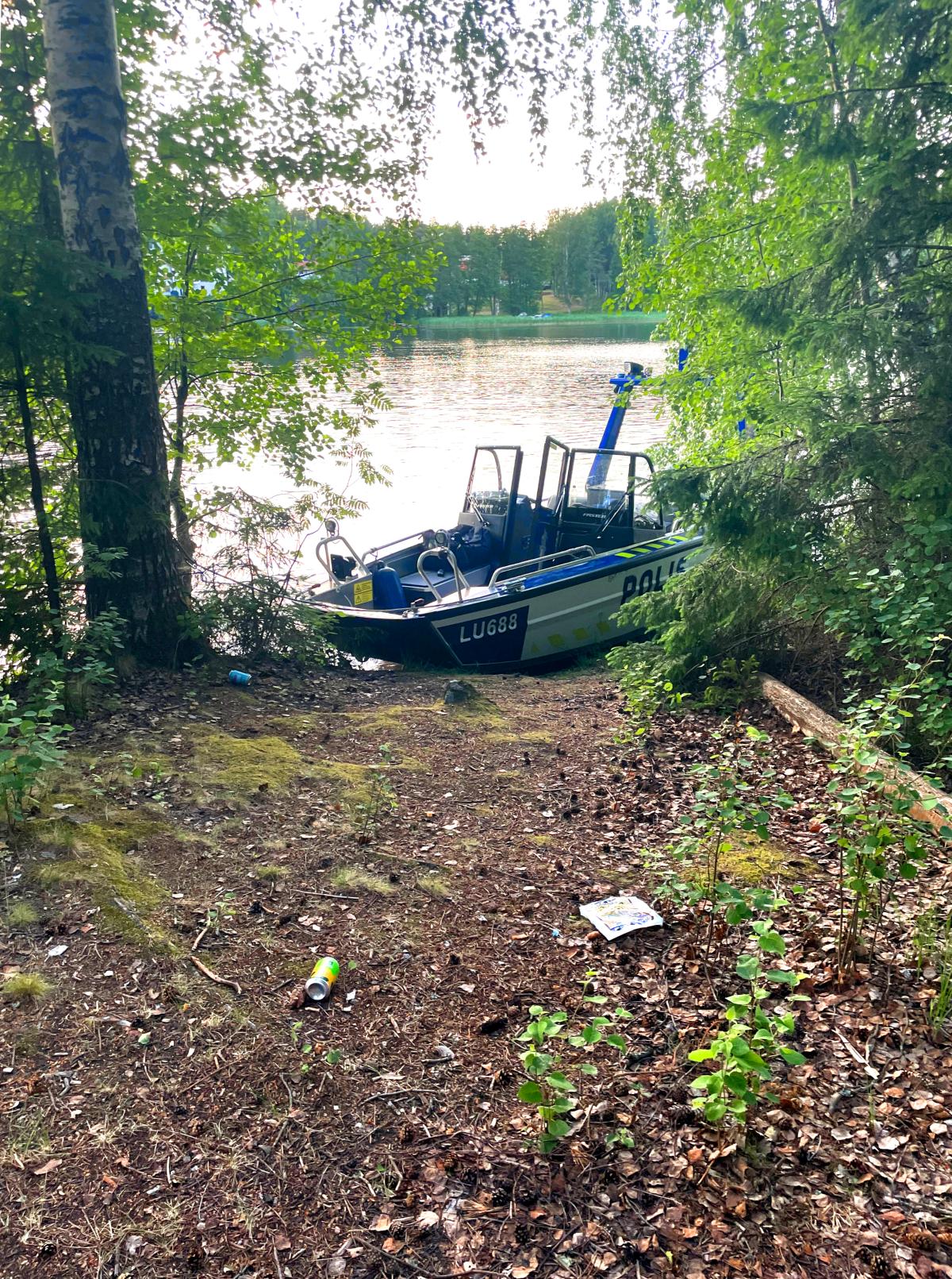 Poliisivene näkyy metsän puiden lomasta järven rannalla. Maassa on roskia.