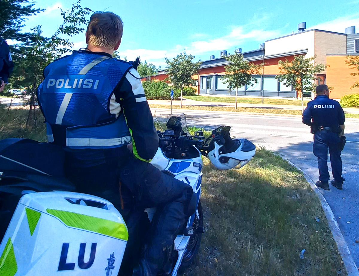 Moottoripyöräpoliisi istuu moottoripyörän päällä ja toinen poliisi valvoo liikennettä.