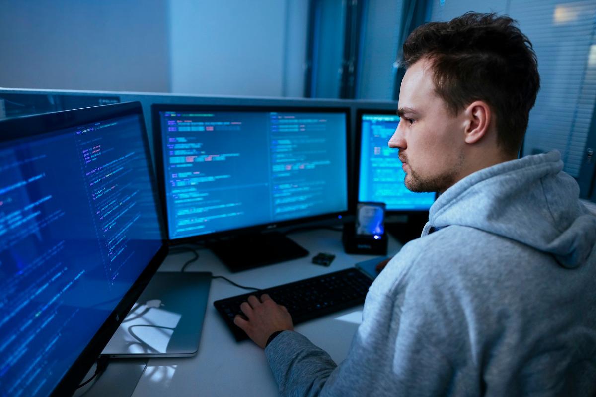 Mies istuu työpöydän ääressä kolmen tietokonenäytön edessä ja tutkii näytöllä näkyvää kyberrikoksiin liittyvää koodia. Kuvituskuva.