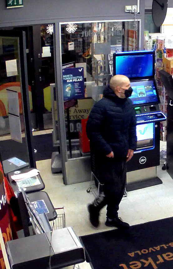 Tummiin vaatteisiin pukeutunut mies saapumassa kauppaan kasvomaski kasvoillaan.